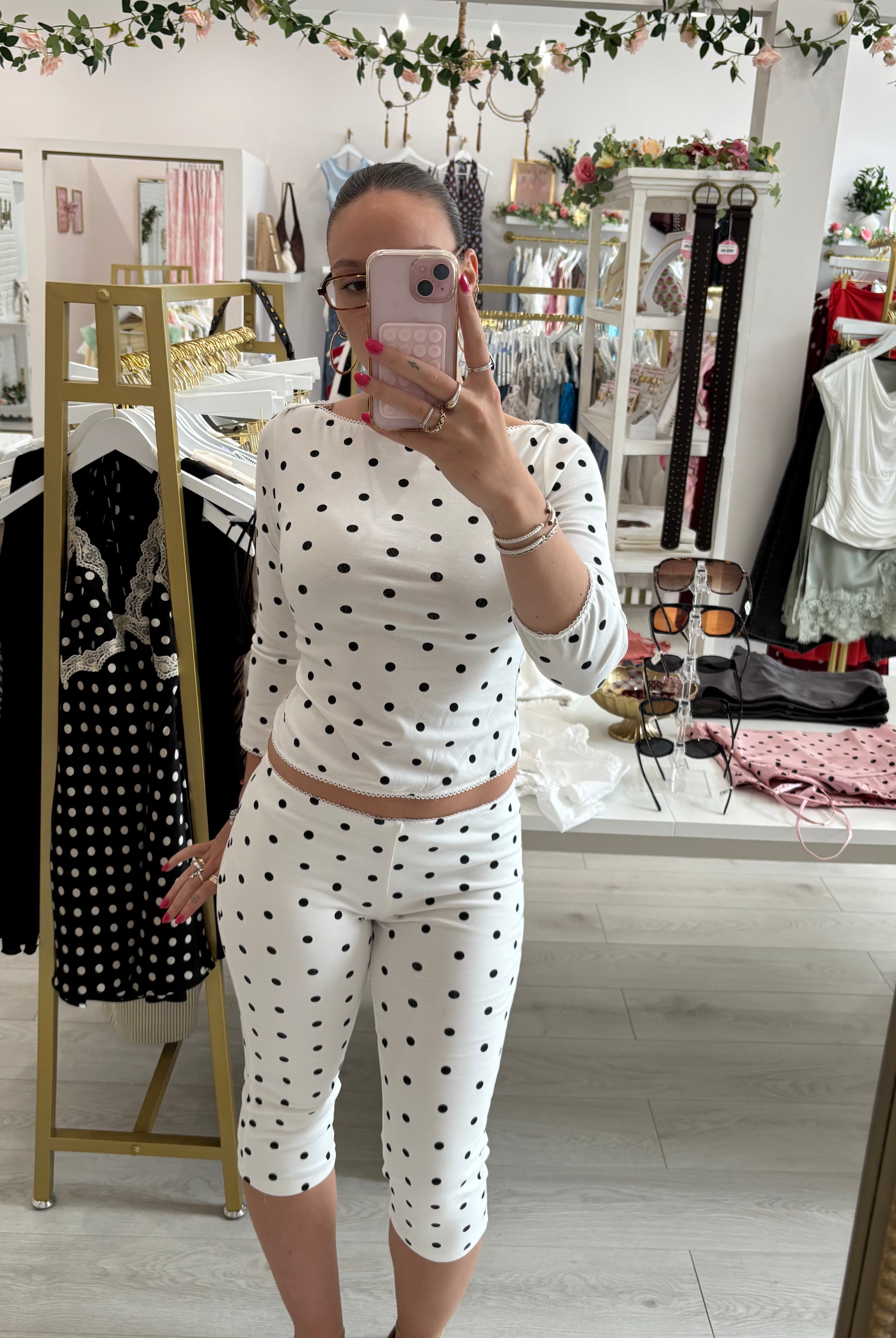 A woman takes a mirror selfie in a boutique showing a matching two-piece outfit consisting of a white long-sleeve top and matching capri pants with black polka dots.