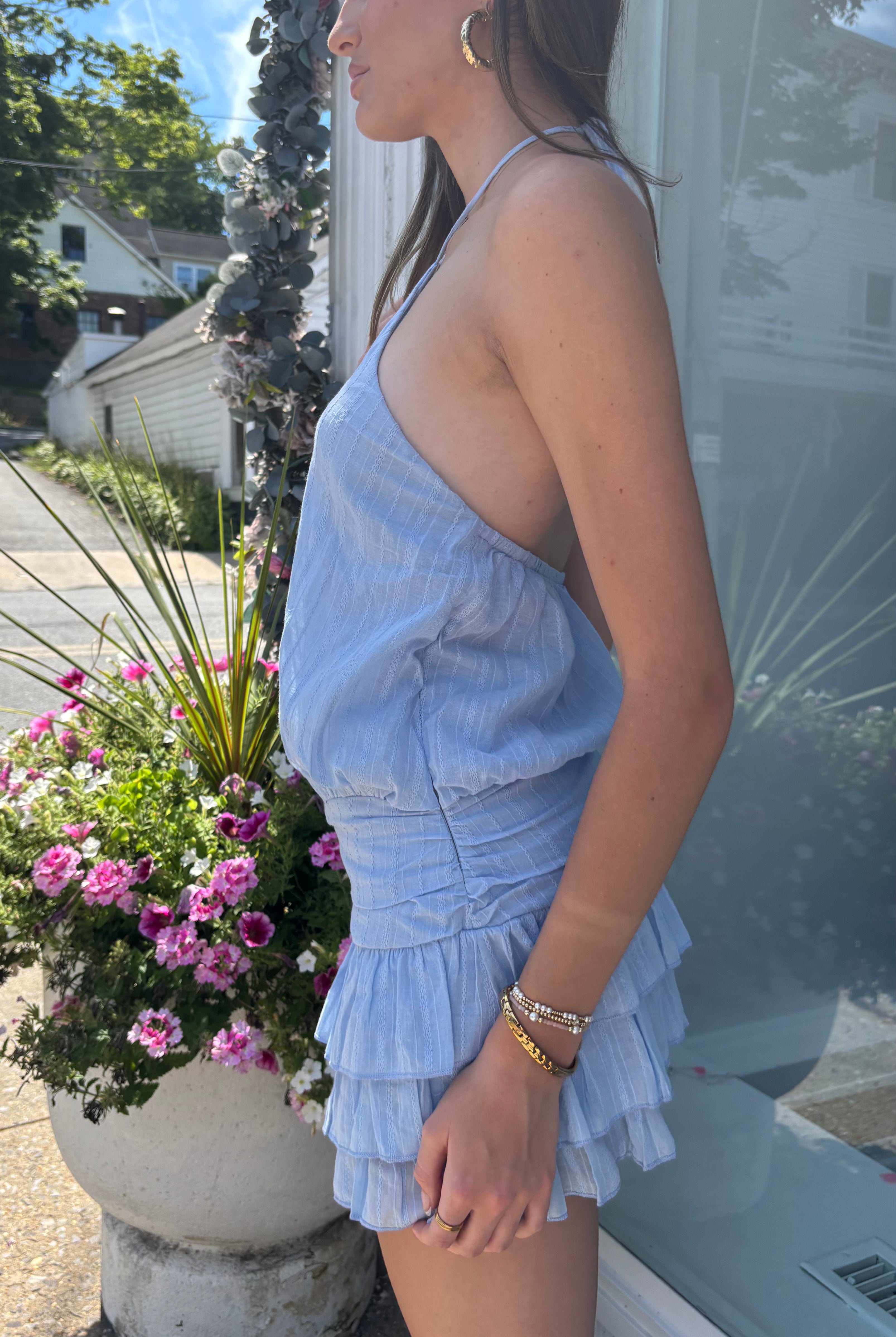A woman wearing a light blue ruffled halter romper stands in profile outdoors next to a storefront window and a planter with pink flowers.