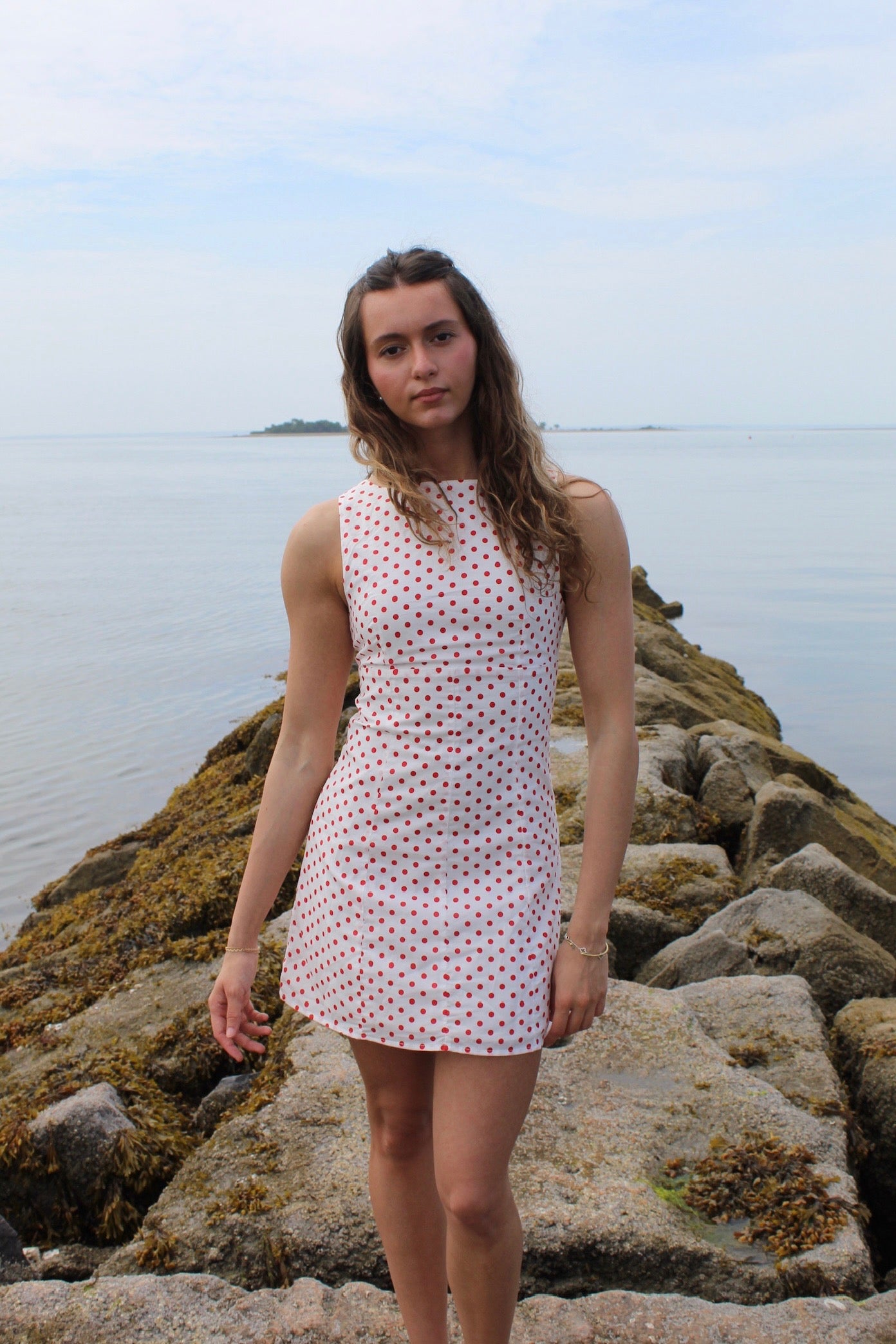 Woman wearing a white mini dress with red polka dots standing on rocks by the water. Fourth of July Mini Dress