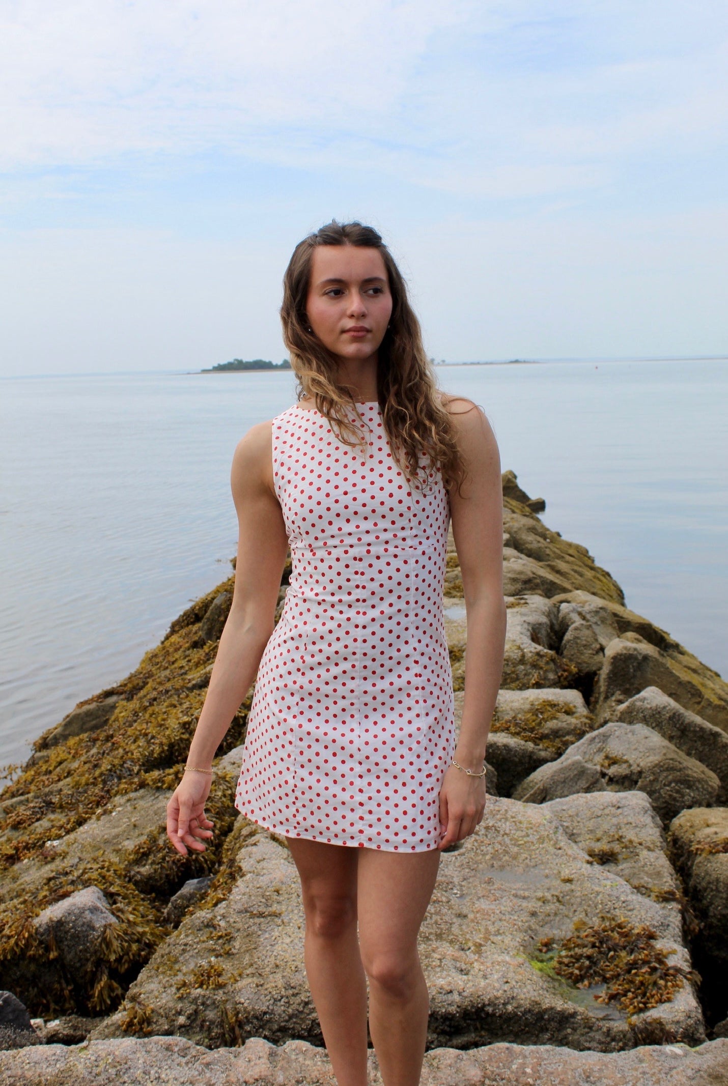 Woman wearing a white mini dress with red polka dots standing on rocks by the water. Fourth of July Mini Dress