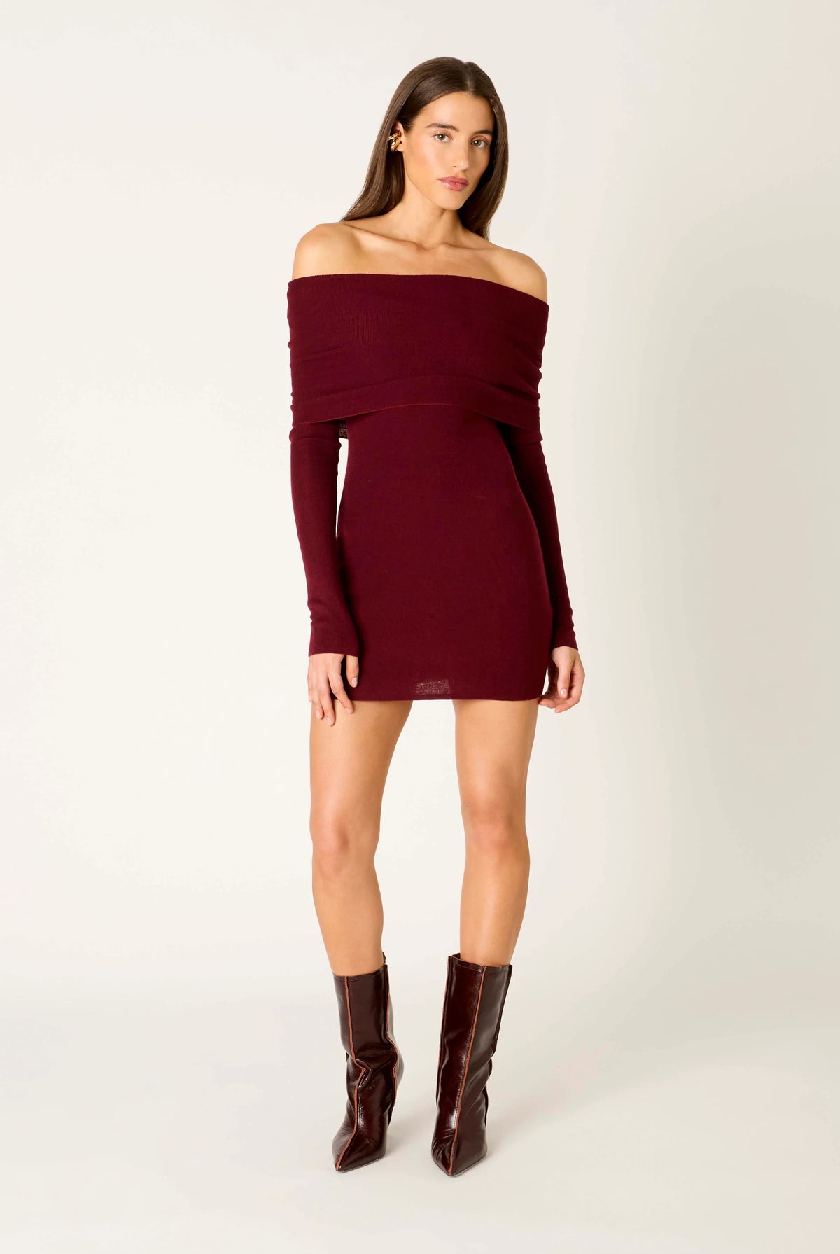A woman modeling an off-the-shoulder long-sleeve mini dress in deep burgundy, paired with dark brown knee-high boots, against a plain light gray studio background.