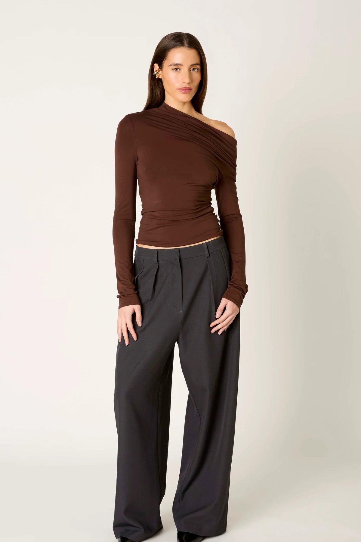 A model wearing a dark brown, asymmetrical one-shoulder long-sleeve top and dark charcoal gray wide-leg trousers, standing against a light gray studio background.