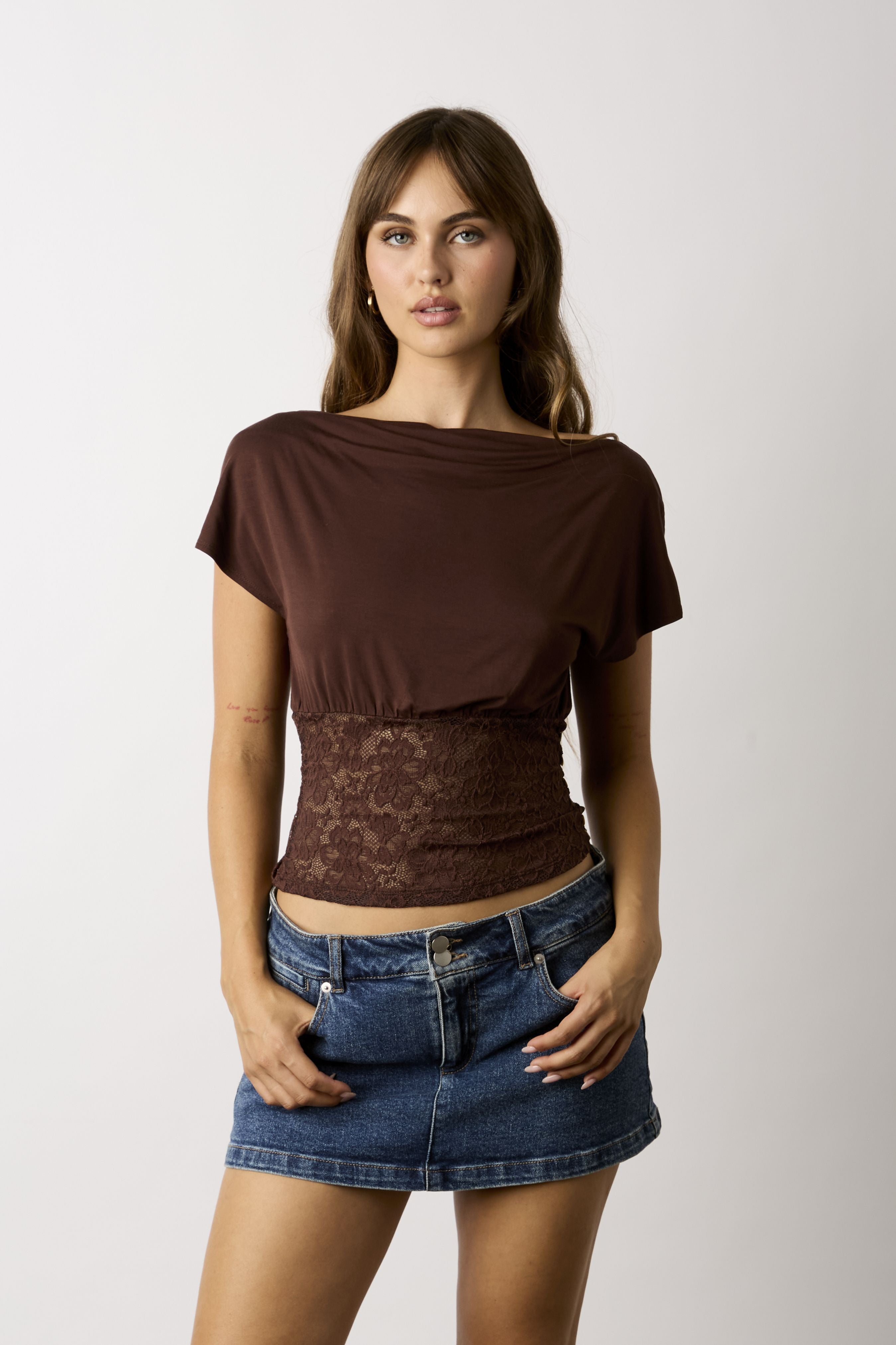 A woman modeling a brown short-sleeve top with a sheer lace waist panel, paired with a blue denim mini skirt, against a plain light gray background.