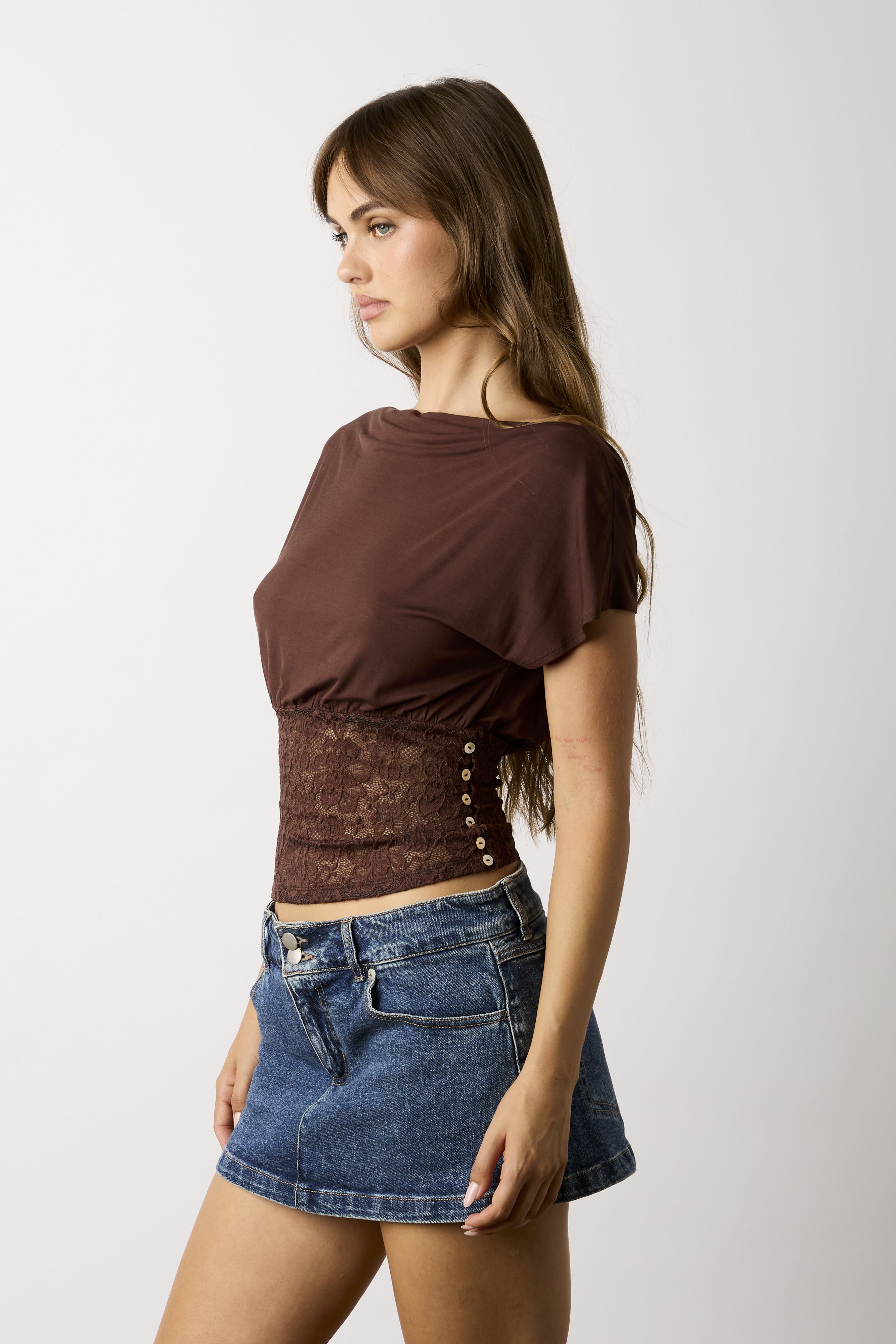 A woman wearing a brown short-sleeve crop top with a lace-up corset-style waist panel and blue denim mini skirt, photographed against a plain light gray studio background.