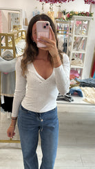 A woman takes a mirror selfie in a boutique, wearing a light gray heathered long-sleeve V-neck top and blue jeans. The boutique has white shelving, clothing racks, and pink tinsel garlands hanging from the ceiling.