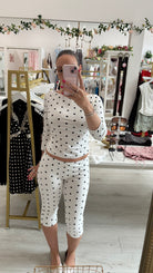 A woman takes a mirror selfie in a boutique showing a matching two-piece outfit consisting of a white long-sleeve top and matching capri pants with black polka dots.