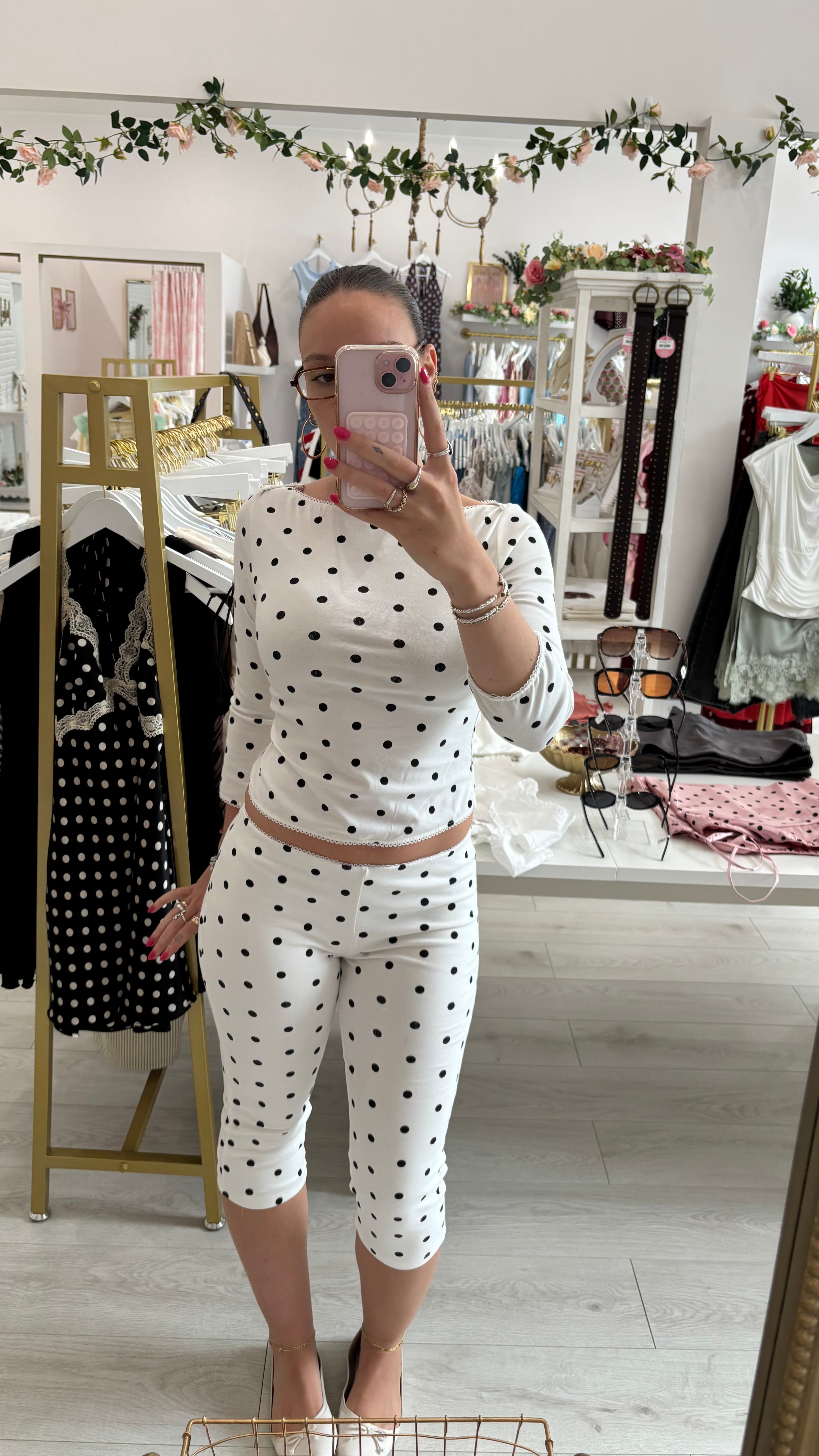A woman takes a mirror selfie in a boutique showing a matching two-piece outfit consisting of a white long-sleeve top and matching capri pants with black polka dots.
