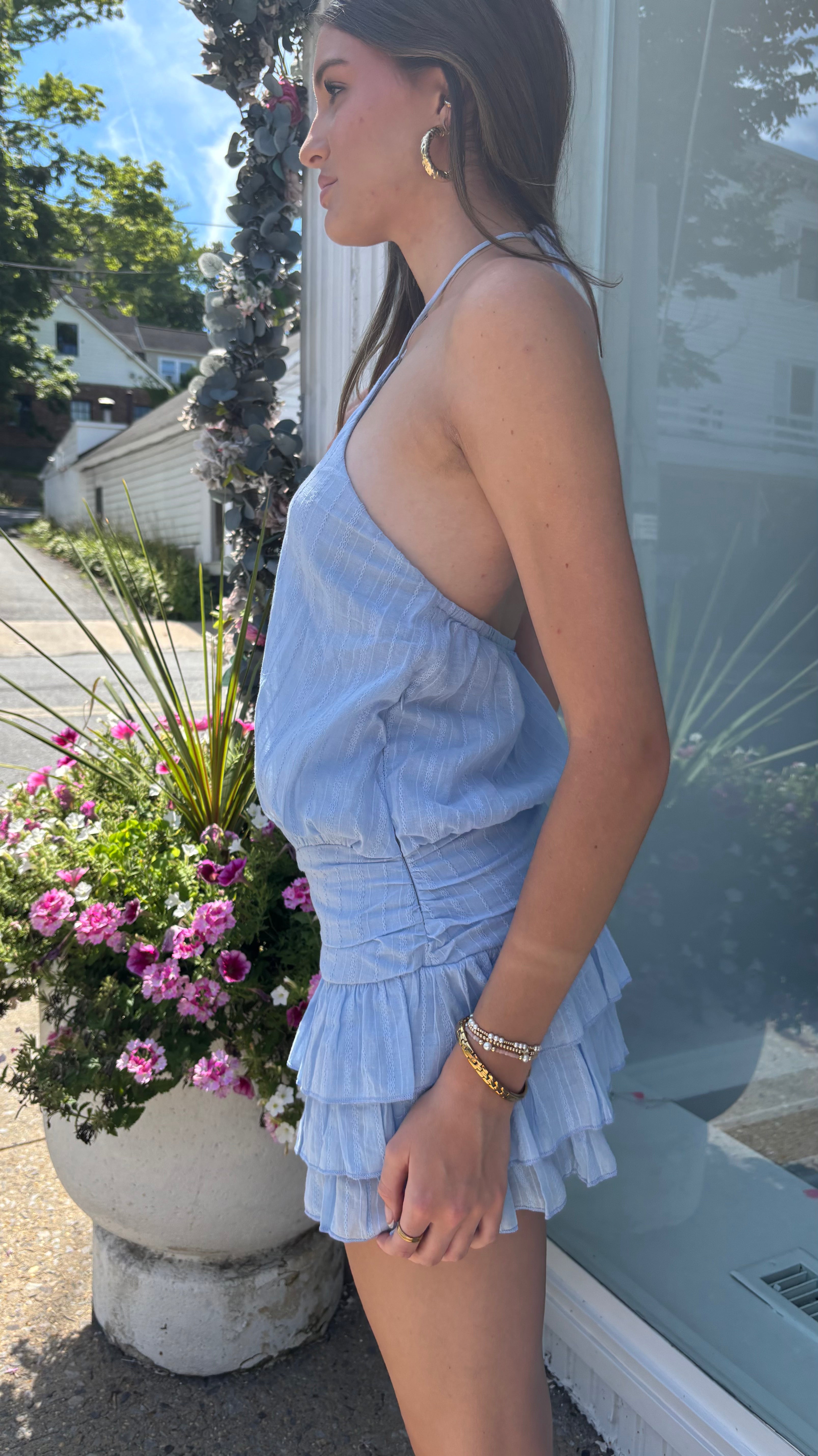 A woman wearing a light blue ruffled halter romper stands in profile outdoors next to a storefront window and a planter with pink flowers.