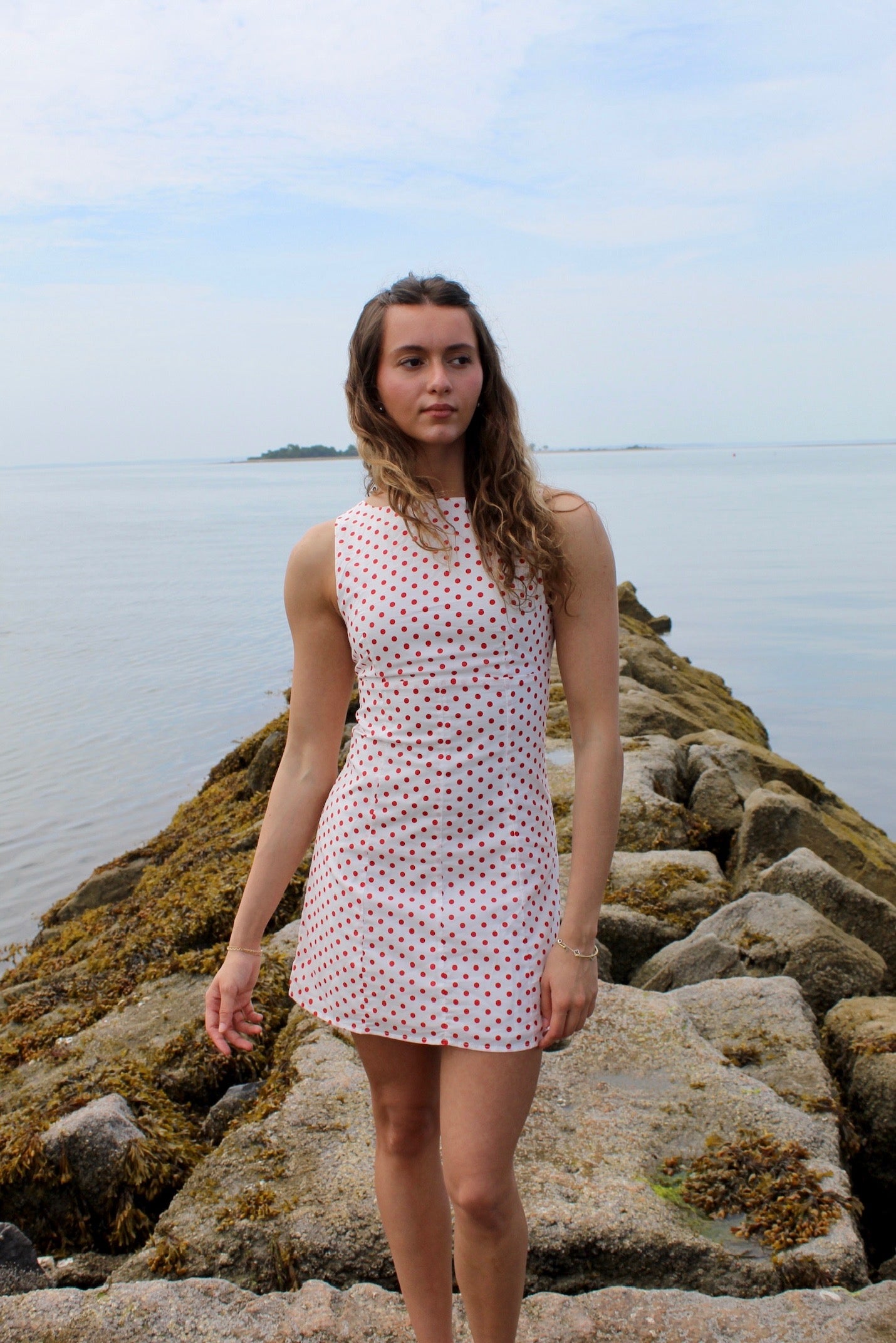 Woman wearing a white mini dress with red polka dots standing on rocks by the water. Fourth of July Mini Dress 