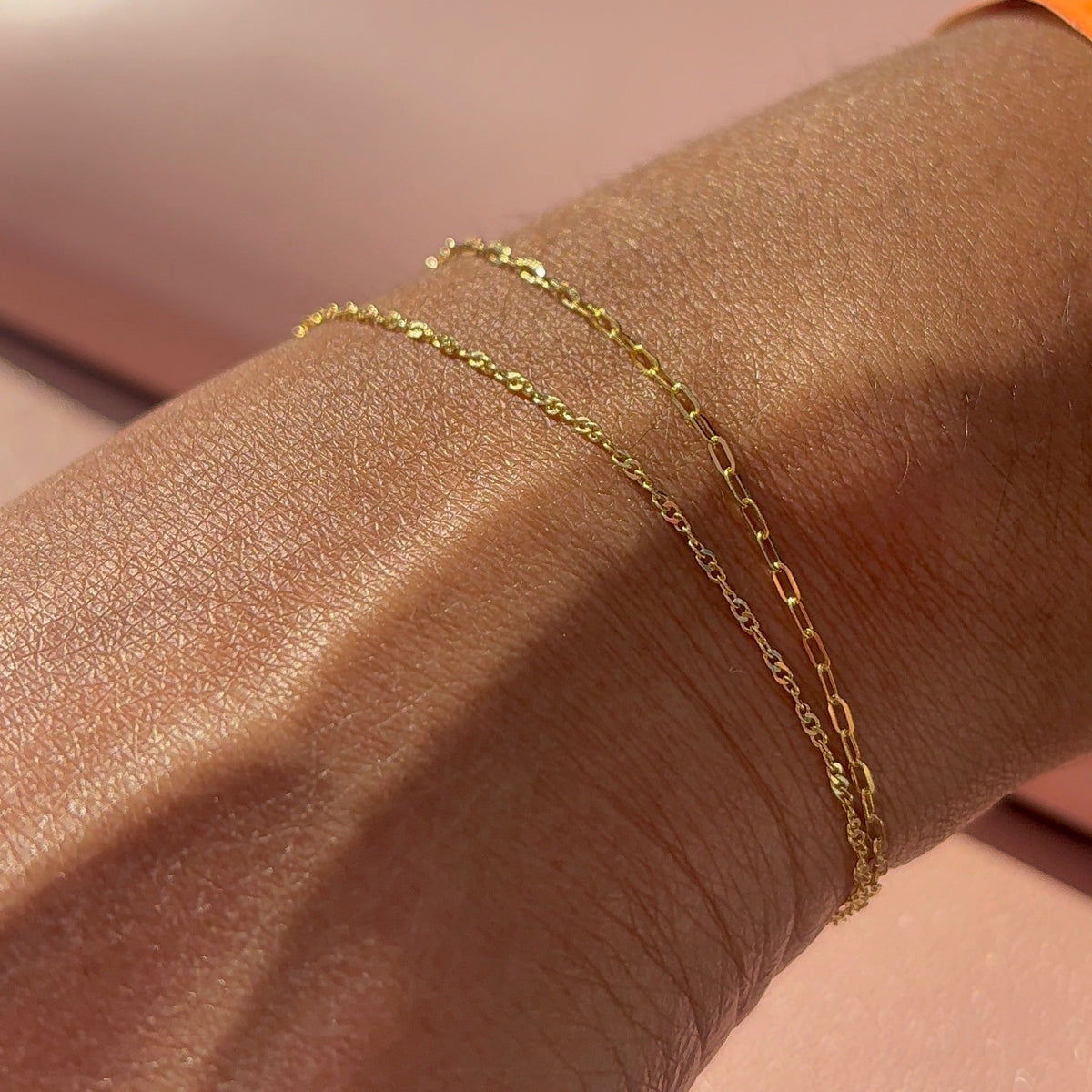 A close-up of a person's forearm wearing two delicate gold chain bracelets, with a pink surface and an orange wristband in the background.