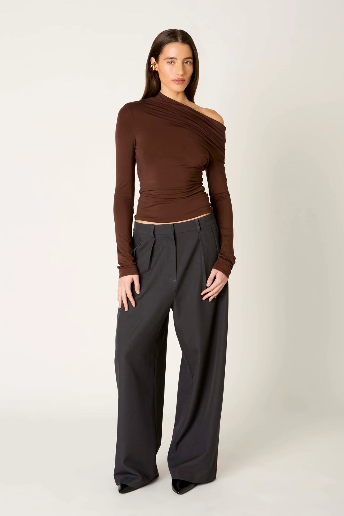 A model wearing a dark brown, asymmetrical one-shoulder long-sleeve top and dark charcoal gray wide-leg trousers, standing against a light gray studio background.