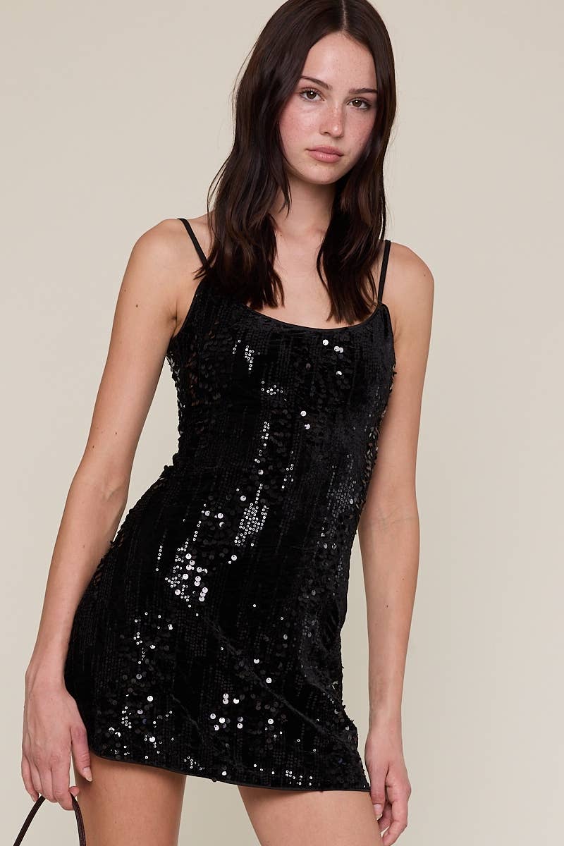A woman modeling a black sequined spaghetti-strap mini dress against a plain beige studio background.