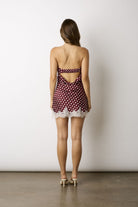 A woman viewed from behind wearing a strapless mini dress with a red and white polka dot pattern, a cutout back, and a white lace hem, standing against a plain white wall.