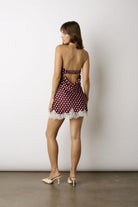 A woman stands with her back to the camera wearing a short burgundy and white polka-dot mini dress with a lace hem and cutout back, paired with white heeled mules, against a plain light gray studio wall.