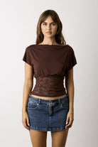 A woman modeling a brown short-sleeve top with a lace waist panel and a blue denim mini skirt, photographed against a light gray studio background.