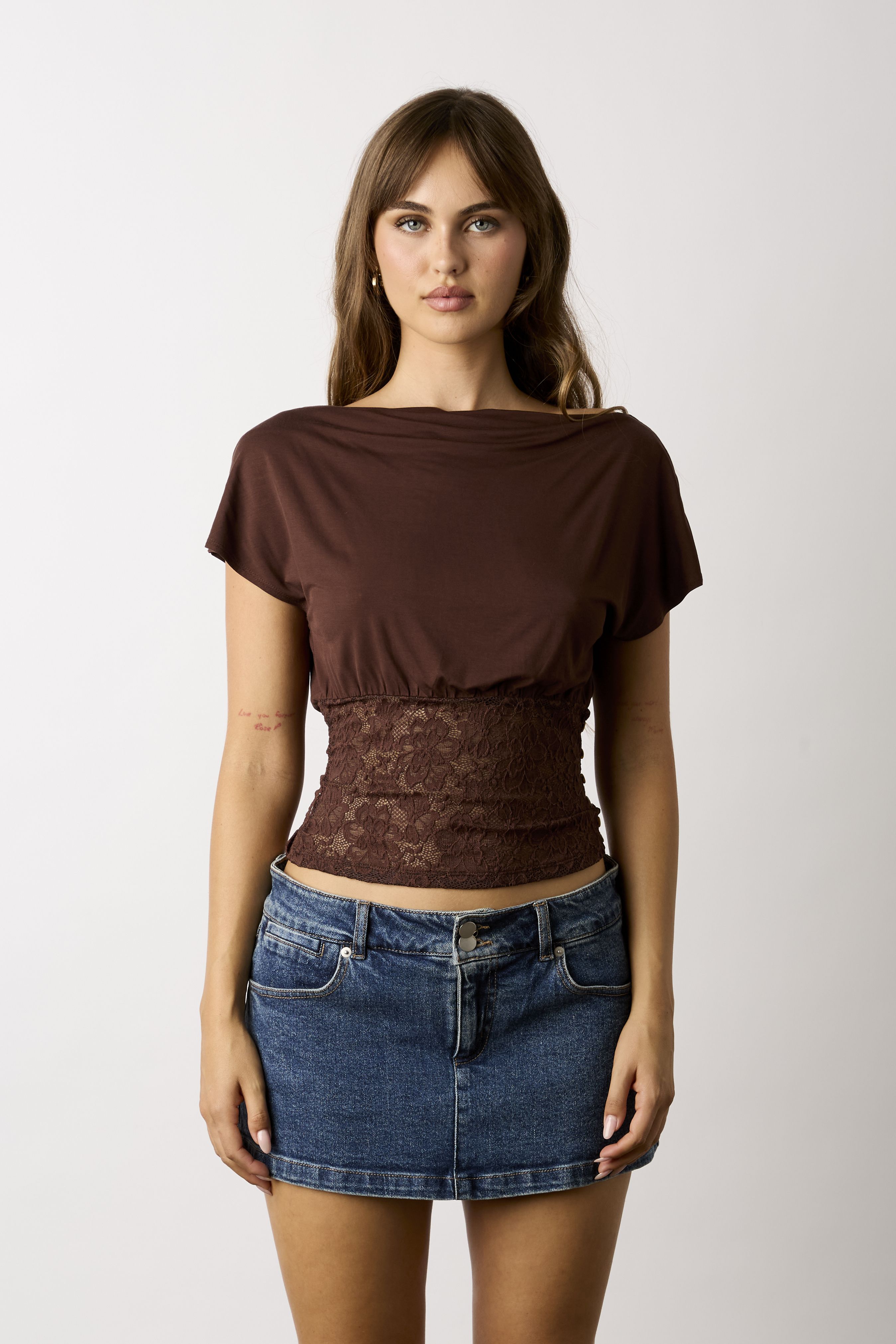 A woman modeling a brown short-sleeve top with a lace waist panel and a blue denim mini skirt, photographed against a light gray studio background.