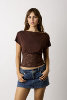 A woman modeling a brown short-sleeve top with a sheer lace waist panel, paired with a blue denim mini skirt, against a plain light gray background.