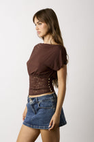 A woman wearing a brown short-sleeve crop top with a lace-up corset-style waist panel and blue denim mini skirt, photographed against a plain light gray studio background.