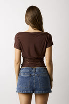 A woman shown from the back wearing a brown short-sleeve top with a wide boat neckline and a ruched waist, paired with blue denim shorts, against a plain light gray background.